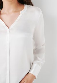 Blusa blanca de seda de mangas largas con escote en V, que presenta una textura suave y un detalle de tapeta de botones.