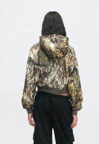 Person med langt, mørkt hår iført en cropped camo-hoodie med barkmønster og sorte løse bukser, stående med ryggen til en hvid baggrund.