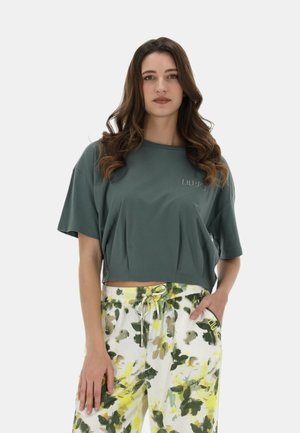 Joven mujer con cabello castaño largo y ondulado, vistiendo una camiseta corta verde oscuro holgada y pantalones blancos con estampado floral verde y amarillo.