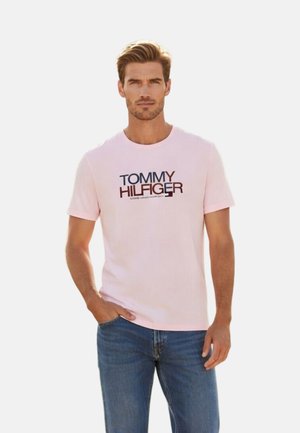 Hombre vistiendo una camiseta rosa claro de Tommy Hilfiger y jeans azules, de pie con una mano en el bolsillo contra un fondo liso.
