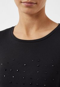Schwarzes Shirt mit runder Ausschnitt und einem strukturierten Muster aus kleinen, glänzenden schwarzen Pailletten, die unregelmäßig über den Stoff verteilt sind.