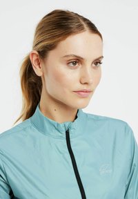 Lyseblå, letvægts cykeljakke med høj krave og frontlynlås. Har et lille logo og en glat tekstur.