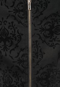Tissu noir avec un motif damassé, une fermeture éclair dorée apparente au centre, contribuant au design détaillé. Finition texturée.