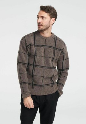 Brązowy sweter wełniany w kratę z czarnymi liniami. Wykończenie z okrągłym dekoltem, ściągacze na rękawach i u dołu. Noszony z ciemnymi spodniami.