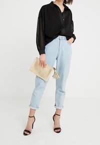 Musta läpikuultava pitkähihaispaita, jossa on pallokuosi, vaaleansininen korkeavyötäröiset farkut, mustat remmikkäät korkokengät ja beige hapsullinen käsilaukku.