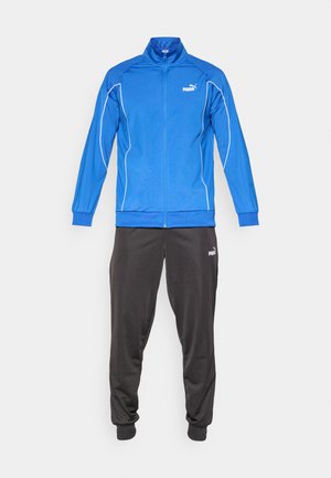 Dres Puma składający się z niebieskiej kurtki zapinanej na zamek z białymi akcentami oraz czarnych spodni dresowych, wykonany z gładkiego materiału z elastycznymi mankietami.