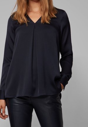 Femme portant un chemisier noir à col en V et manches longues avec un pli à l'avant et un pantalon en cuir noir, debout devant un fond gris.