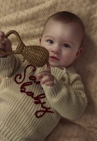 Maglione lavorato a maglia beige con il testo ricamato in rosso "baby" e bottoni in legno. Bambino che tiene un giocattolo intrecciato con un manico, su uno sfondo beige texturizzato.