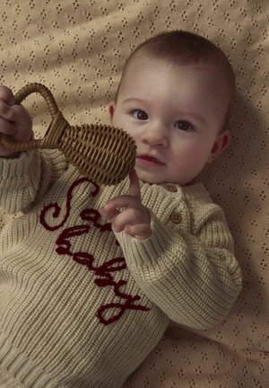 Pull en tricot beige avec texte brodé en rouge "baby" et boutons en bois. Un nourrisson tenant un jouet tissé avec une poignée, sur un fond beige texturé.