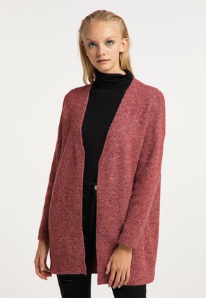 Frau trägt eine lange, offene rote Strickjacke über einem schwarzen Rollkragenpullover und schwarzen Hosen und steht vor einem einfarbigen hellen Hintergrund.
