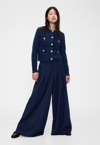 Tamsiai mėlynas vilnonis kardiganas su auksiniais mygtukais ir dviem krūtinės kišenėmis, derinamas su plačiomis tamsiai mėlynomis dryžuotomis kelnėmis ir juodais bateliais su raišteliais.