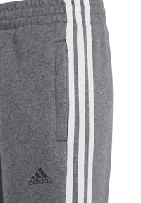 Grå fleece-joggers med vita sidostreck och en sidficka. Har en svart Adidas-logotyp nära midjan. Mjuk textur.