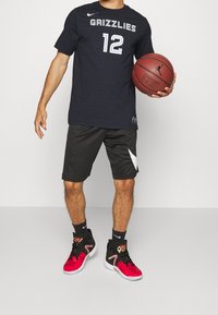 Černý basketbalový dres s nápisem "GRIZZLIES" a číslem "12" v bílé barvě, doplněný o černé šortky s bílým akcentem a černým a červeným sportovním obuvím.