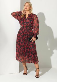 Czerwona sukienka w kwiatowy wzór, wykonana z przezroczystego materiału, z marszczonym pasem, długimi bufkami i zwiewnym dołem. Modelka trzyma czarną kopertówkę.