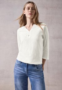 Weißes Langarmoberteil mit einem Reverskragen, kombiniert mit blauen Jeans. Der Stoff wirkt weich und hat eine lockere Passform.