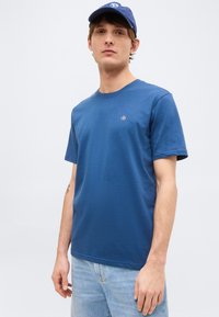 Νέος άνδρας με μπλε καπέλο, μπλε T-shirt με μικρό λογότυπο στο στήθος και ανοιχτό μπλε τζιν, στέκεται μπροστά σε απλό ανοιχτό φόντο.