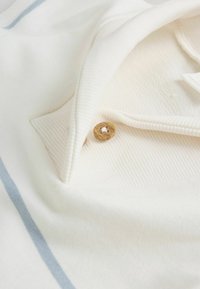 Bela polo majica z izrezljanim ovratnikom, ki ima eno leseno gumb in svetlo modro črto blizu roba. Mehka tekstura tkanine.