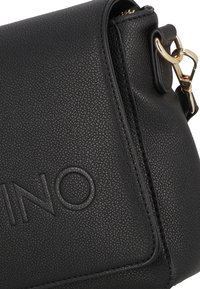 Borsa a tracolla in pelle nera con texture, caratterizzata da una clip in metallo dorato e branding in rilievo. Forma compatta con chiusura a zip.