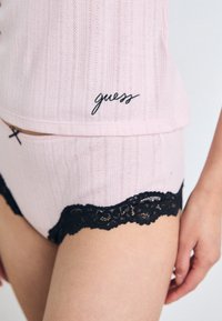 Tessuto rosa a coste con pizzo nero decorativo sulla parte inferiore. La parte superiore presenta un logo "guess" ricamato in nero. Design semplice con tocchi giocosi.
