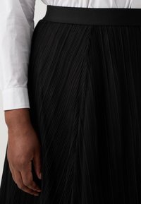 Svart plisserad kjol med en slät tyg och ett brett, matt midjeband. Handen vilar på kjolen och visar upp textur och designdetalj.