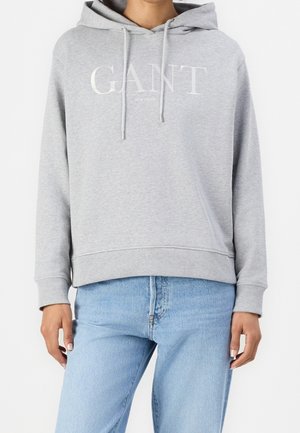 Kobieta ubrana w jasnoszarą bluzę z kapturem GANT z troczkami i jasnoniebieskie dżinsy, pokazana od ramion do bioder na białym tle.