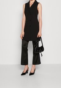 Schwarze ärmellose Blazer mit doppelten Knöpfen, kombiniert mit schwarzen satinierten ausgestellt geschnittenen Hosen. Accessoiert mit einer kleinen schwarzen Tasche mit runden Beschlägen.