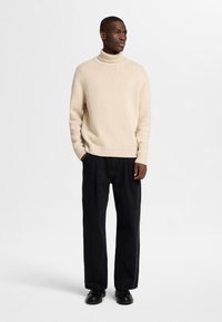 Bež pletena pulover s turtleneck-om in rebrastimi manšetami, v kombinaciji z ohlapnimi črnimi hlačami in temnimi čevlji. Gladka tekstura, preprost dizajn.