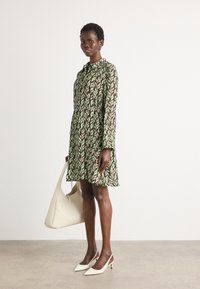 Abito a maniche lunghe e colletto, con una fantasia verde e marrone, abbinato a una borsa crema e a delle décolleté bianche con punta affusolata.