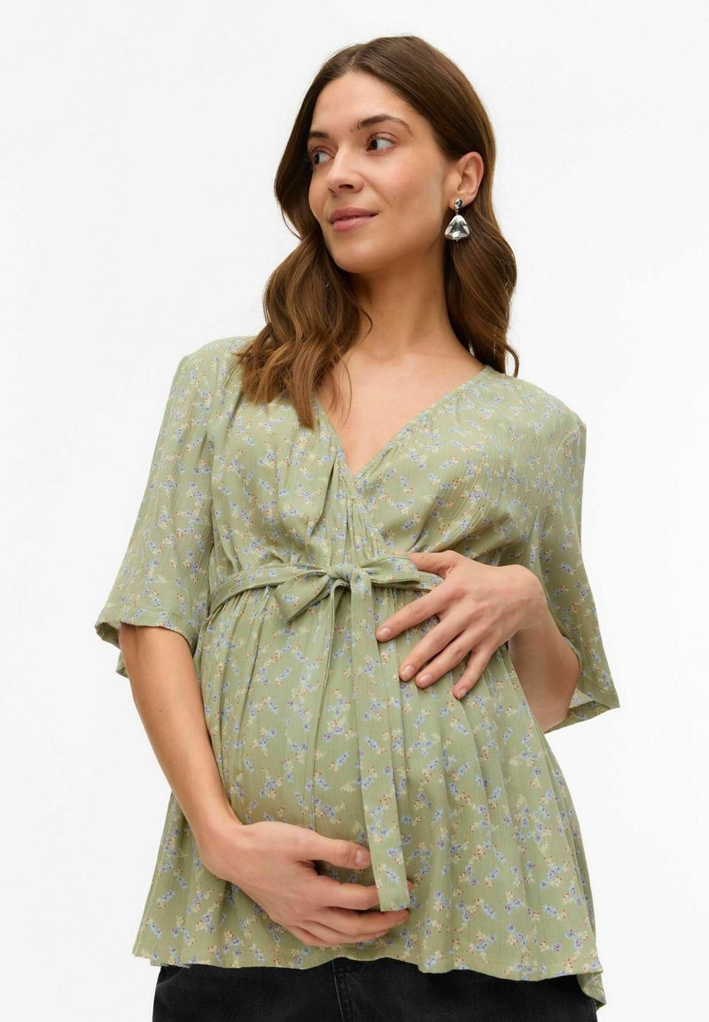 Zelena cvetna oblačila za nosečnice s kratkimi rokavi, V-izrezom, z vezavo spredaj in mehko, strukturirano tkanino. Ima ohlapen kroj, ki ustreza rastočemu trebuhu.