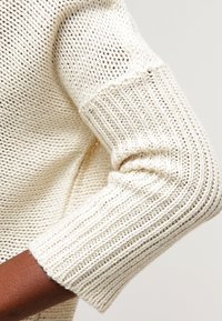 Pletený svetr v krémové barvě s volným vzorem a žebrovanými manžetami na rukávech. Kompozice zdůrazňuje texturovanou látku a volný střih.