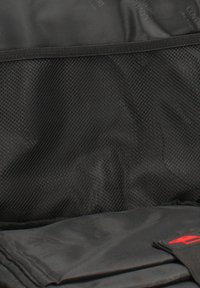 Poche en mesh noire et organiseur en tissu à l'intérieur d'un sac, avec un rabat plié présentant un logo ou un élément de design rouge.