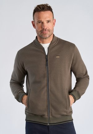 Man wearing a brown zip-up jacket with hands in pockets, standing against a plain light background.