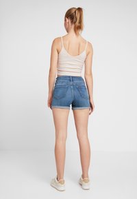 Denim shorts in medium blue with a frayed hem, two back pockets, worn with a light pink ribbed tank top and beige sneakers.