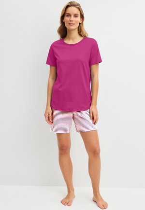 Vrouw staat op blote voeten, draagt een felroze T-shirt met korte mouwen en witte korte broek met dunne rode horizontale strepen tegen een effen achtergrond.