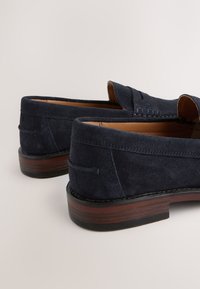 Marineblaue Wildleder-Loafer mit abgerundetem Zehenbereich, gesteppten Details und einer braunen Holzsohle. Glatte Textur und zum Hineinschlüpfen.