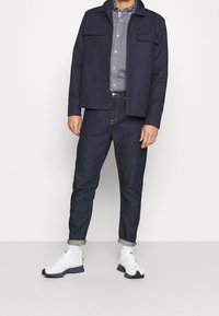 Giacca blu navy con chiusura a bottoni, due tasche sul petto; camicia azzurro chiaro con bottoni; jeans blu scuro; sneaker bianche e nere; maniche arrotolate.