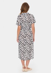 Długa, luźno dopasowana sukienka w czarno-białym wzorze liści, z krótkimi rękawami i detalem związania w talii. Miękka, lekka tkanina.