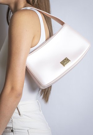 Femme portant un haut blanc sans manches et un pantalon, portant un sac à main rose pâle avec une plaque logo dorée sur son épaule.