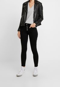 Jeans skinny neri a vita alta con cinque bottoni anteriori, abbinati a una t-shirt grigia a V e una giacca da moto in pelle nera. Sneakers bianche.