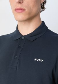 Tmavomodrá polo košeľa s textúrovanou látkou, so golierom, tromi gombíkmi a bielym logom "HUGO" na ľavej hrudi.