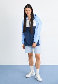 Coupe-vent bleu clair et marine avec un panneau blanc ; présente une fermeture éclair à l'avant, une capuche et des poignets élastiques ; associé à un short bleu clair et des baskets blanches.