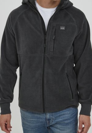 Mann trägt eine dunkelgraue Fleecejacke mit Reißverschluss, Brust- und Seitentaschen, über weißem Hemd und blauen Jeans.