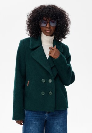 Kobieta z kręconymi włosami, ubrana w przesadnie duże okulary przeciwsłoneczne, ciemnozielony płaszcz dwurzędowy, biały golf oraz niebieskie dżinsy.