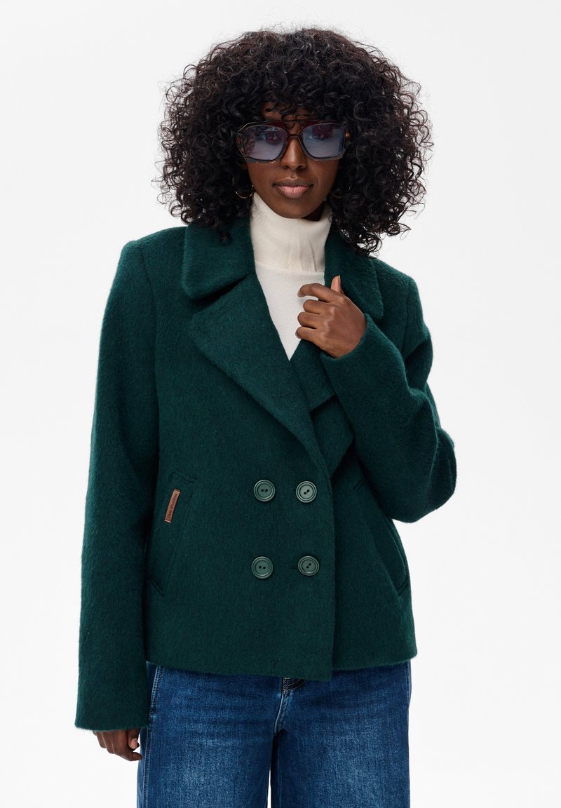 Kobieta z kręconymi włosami, ubrana w przesadnie duże okulary przeciwsłoneczne, ciemnozielony płaszcz dwurzędowy, biały golf oraz niebieskie dżinsy.