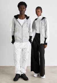 Uomo e donna che indossano felpe grigie con zip abbinate, con accenti in bianco e nero, in piedi all'interno su un pavimento beige contro un muro bianco.
