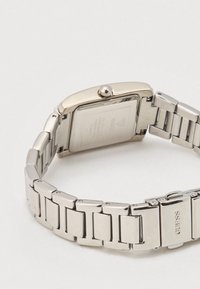 Srebrni ručni sat od nehrđajućeg čelika s pravokutnim brojčanikom, poliranim okvirom i povezanim remenom; ima krunu i ugravirani logotip marke na kopči.