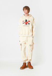 Joven de pie vistiendo una sudadera crema con un gráfico rojo y negro, pantalones cargo a juego y botas de cordones marrones, sobre un fondo blanco liso.