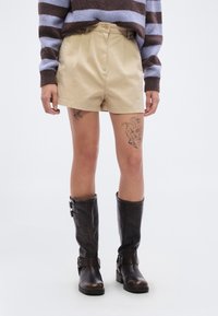 Μπεζ βαμβακερά σορτς με πιέτες, σε συνδυασμό με σκούρα καφέ δερμάτινες μπότες μέχρι το γόνατο με αγκράφες. Ριγέ πλεκτό πουλόβερ σε καφέ και μπλε.