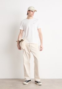 Hvit T-skjorte, kremfargede bukser, svarte joggesko. Tilbehør inkluderer en beige cap og en beige jakke holdt i den ene hånden. Enkelt, uformelt antrekk.