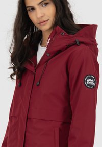 Rode waterdichte jas met capuchon, voorzien van een rits, trekkoorden en een ronde logo patch op de mouw. Gladde textuur en praktische zakken.